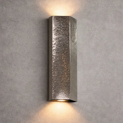 A modern rectangular wall sconce with a hammered metallic finish, mounted on a textured gray concrete wall and casting a warm light upward and downward.