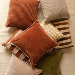 A styled collection of home decor throw pillows in various textures and patterns, including rust orange corduroy, beige ribbed velvet, striped velvet, and olive green velvet, arranged on a light wood floor.
