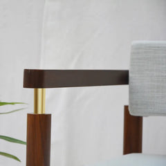 Close-up of a modern wooden chair with a dark brown wood frame, a gold metal accent post, and a light gray textured fabric seat against a white draped background.