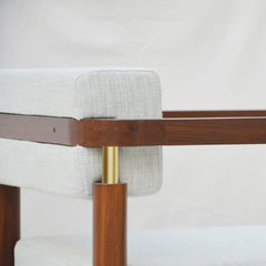 Close-up of a modern upholstered chair or bench corner showing light gray fabric, a dark brown wooden frame, and a brass metal support against a plain light background.