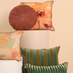 A styled collection of decorative throw pillows displayed on white pedestals against a warm beige wall. The set includes a round brown pleated cushion, a square floral print cushion, a green and gold striped cushion, and a green and white striped cushion.