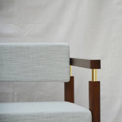 Close-up of a modern armchair corner showing a light gray textured fabric seat and back cushion with dark brown wooden arms and legs accented by gold metal spacers, set against a draped white fabric backdrop.