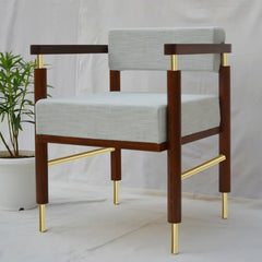 A modern armchair with a dark brown wooden frame, light gray textured fabric upholstery, and polished gold metal accents on the legs and arm supports, set against a white fabric backdrop with a potted plant on the left.