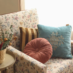 A cozy living room scene featuring a floral armchair with decorative throw pillows, including a round dusty rose tufted cushion, a teal floral pillow, and a brown-and-gold striped pillow.