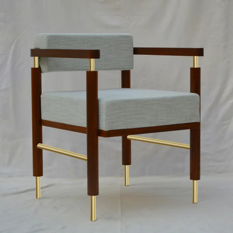 A modern armchair with a dark brown wooden frame, light gray textured fabric cushions, and polished gold metal accents on the legs and armrests, set against a neutral draped backdrop.