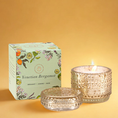Venetian Bergamot Fragrance with Trinket Jar Candle, lit gold patterned glass jar and lid beside box on warm yellow