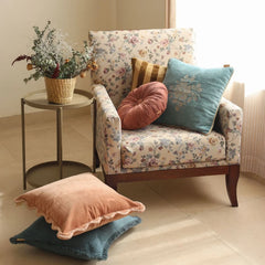 A floral armchair in a bright room, styled with a teal floral throw pillow, a dusty rose velvet round pillow, and a peach ruffled throw pillow. A small round side table with a wicker basket of flowers sits to the left.