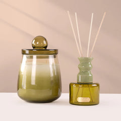 Cosmic Set - A glass jar candle with a matching lid and a reed diffuser bottle with reeds, both in olive green glass, displayed on a white surface against a beige background.