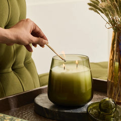 A olive green glass jar candle with two wicks is lit with a wooden match by a person. The candle sits on a dark stone tray next to a matching glass candle lid.