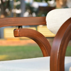 Close-up of a wooden outdoor chair with a curved armrest, a cream-colored fabric cushion, and a blurred garden background.