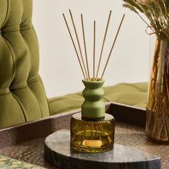 A reed diffuser with a light green ceramic bottle and natural wood reeds, set on a dark tray next to a tufted green chair and a glass vase. 