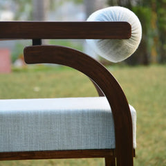 A close-up of a modern wooden chair with a curved dark brown frame, a light blue textured seat cushion, and a round white knit headrest pillow, set outdoors on a grassy lawn.