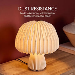 A mushroom-shaped table lamp with a pleated paper shade and base, glowing warm white on a wooden table against a dark red-brown wall. White text at the top reads 'DUST RESISTANCE' with smaller descriptive copy.