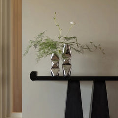 Two metallic silver faceted ceramic vases, chrome dented design with green foliage and white flowers, displayed on a black console table against a beige wall.