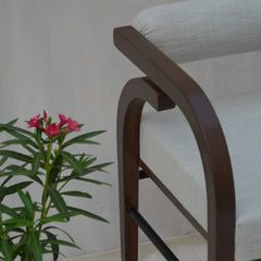 A close-up of a modern armchair with a dark brown wooden frame and light beige textured fabric upholstery, next to a green plant with pink flowers against a neutral light gray wall.