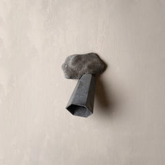 A sculptural wall-mounted light fixture shaped like a dark gray rock with a faceted geometric metal tube extending downward, set against a textured beige plaster wall.