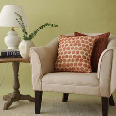 A beige upholstered armchair with two decorative throw pillows, one featuring a rust-orange polka dot pattern and the other a solid dark reddish-brown, styled in a room setting next to a wooden side table with a lamp and books against a sage green wall.