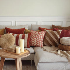 A styled living room scene featuring a light beige sectional sofa heavily decorated with multiple textured throw pillows in rust red, terracotta, beige, and brown patterns, alongside a matching fringed throw blanket and Cuddle Sand Velvet Round Cushion.