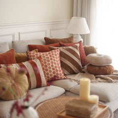 A cozy living room scene with a light beige sofa styled with multiple decorative throw pillows in rust, orange, brown, cream, and striped patterns. A white table lamp sits on a side table next to the sofa, and a round tray with candles is in the foreground.