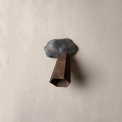A rustic metal wall hook shaped like a dark gray rock with a rusted brown cone below it, mounted on a textured beige plaster wall.