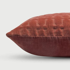 A close-up of a textured, quilted throw pillow in a rust red velvet fabric, shown against a light gray background.