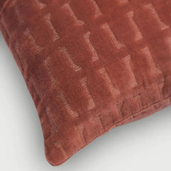 A close-up of a textured throw pillow in a dusty terracotta rust color, featuring a subtle woven geometric pattern, shown on a light gray background.