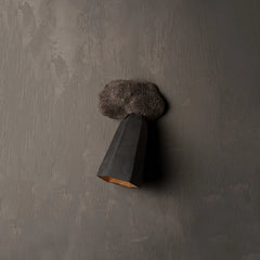 A modern wall sconce with a dark charcoal gray geometric cone shade and a fluffy dark gray faux-fur cloud-like top, mounted on a textured gray plaster wall.