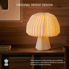 A mushroom origami table lamp with a pleated, accordion-style shade and base, glowing with a warm yellow light. It is placed on a dark wooden table against a matching wood-paneled wall.