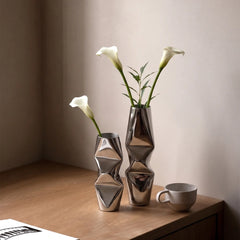 Two Chrome Dented metallic silver geometric flower vases with white calla lilies on a wooden table, alongside a small speckled ceramic cup against a warm beige wall.