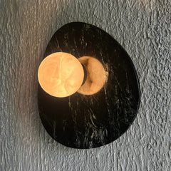 Aureole Green Textured Wall Sconce featuring a polished alabaster black stone base with a glowing warm yellow-orange circular light element, mounted on a textured gray plaster wall.