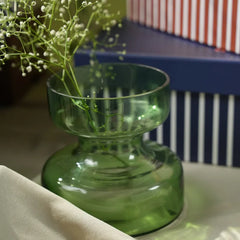 A green glass vase with a flared rim and ribbed neck, set on a white surface with a striped blue and white box in the background.