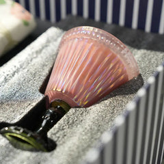 A pink iridescent glass candle with a flared rim, nestled in gray foam packaging inside a striped box.
