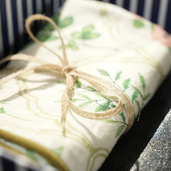 A close-up of a rolled cream-colored Cushion Cover, printed with green botanical motifs and tied with a natural jute twine bow. The background shows a dark navy and white striped gift box.