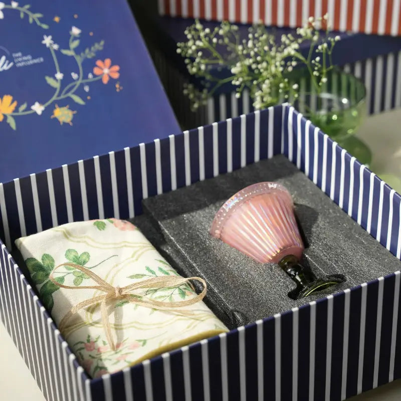 A Cushion Cover and a pink Votive Candle with a flared rim, nestled in gray foam packaging inside a striped box.