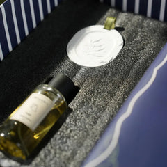 A small clear glass bottle filled with room spray next to a white diffusion stone with an embossed botanical design. Presented inside a blue-and-white striped gift box.