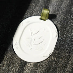 A white diffusion stone with an embossed leaf design and a small olive green ribbon loop, resting on a textured black surface.