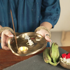 Taral Puja Thaali held in both hands with lit diya and incense beside raw bananas, antique brass finish, front view
