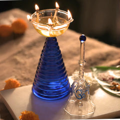 Glass Tall Diya & Ringing Bell Blue, lit diya with oil and glass bell on white tray with marigolds
