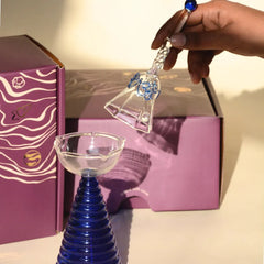 Glass Tall Diya & Ringing Bell Blue, hand lifting glass bell above cobalt blue diya, gift boxes behind