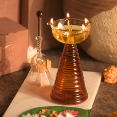 Glass Tall Diya & Ringing Bell Amber, lit diya with oil and glass bell on white tray with marigolds