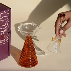 Glass Tall Diya & Ringing Bell Amber, hand holding clear glass bell with gold Om detail beside amber diya