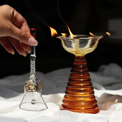 Glass Tall Diya & Ringing Bell Amber, hand ringing glass bell beside lit diya on dark background