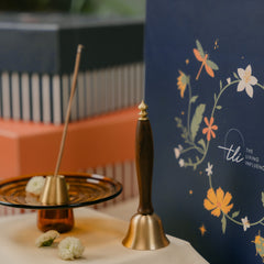 A brass bell with a dark wooden handle  next to a decorative amber glass incense holder along a navy blue gift box with floral illustrations.