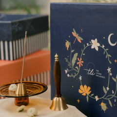 A brass bell, a brass incense holder with a smoking incense stick next to a navy blue decorative box featuring floral and moon illustrations.