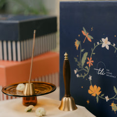 A brass bell with a dark wooden handle stands on a table next to a glass incense holder with a incense stick. 