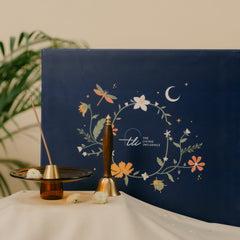 Brass and Natural Wood Bell and brass incense holder, a reed diffuser next to a navy blue gift box with floral illustrations.