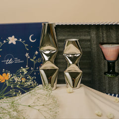 Two metallic silver geometric vases with twisted design, displayed on a draped cream fabric surface alongside a blue decorative box and a pink scented candle in a dark glass holder.