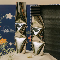 Two modern chrome geometric vases with a faceted, folded design, displayed on a white surface with a blue floral gift box in the background.