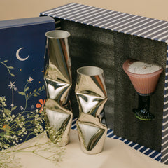 A gift set containing two metallic chrome dented vases and a pink glass scented candle holder, presented in a striped box with a navy blue floral gift box on a beige surface.