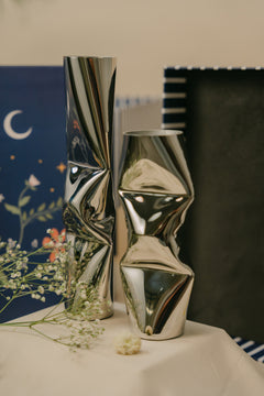 Two modern metallic chrome finish vases with sculptural, twisted geometric shapes, displayed on a draped cream tablecloth.